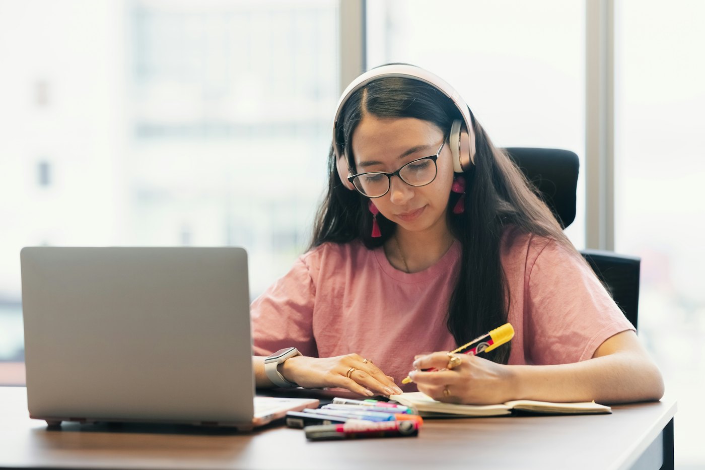 A high school learner in headphones takes notes during an online lesson on her laptop